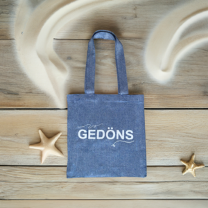 Blaue Stofftasche mit dem weißen Schriftzug „Gedöns“ vor einem Holzuntergrund. Neben der Tasche liegen zwei dekorative Seesterne, die eine sommerliche, maritime Atmosphäre vermitteln.