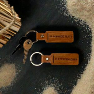 Zwei Holz-Schl&uuml;sselanh&auml;nger mit Gravuren &bdquo;Ik snack Platt&ldquo; und &bdquo;Plattschnacker&ldquo;, auf dunklem Hintergrund mit Sandk&ouml;rnern, daneben ein Schl&uuml;sselbund und Holzst&auml;be.