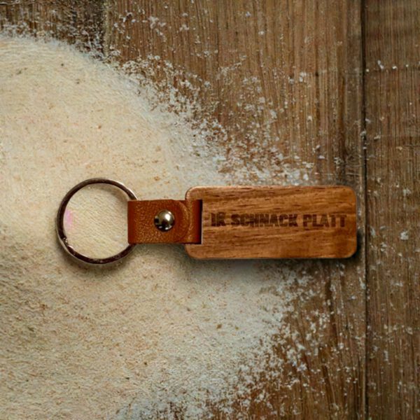 Plattdeutscher Schlüsselanhänger aus Buchenholz mit Gravur „Ik snack Platt“ und Lederschlaufe, auf einer sandbestäubten Holzfläche platziert.